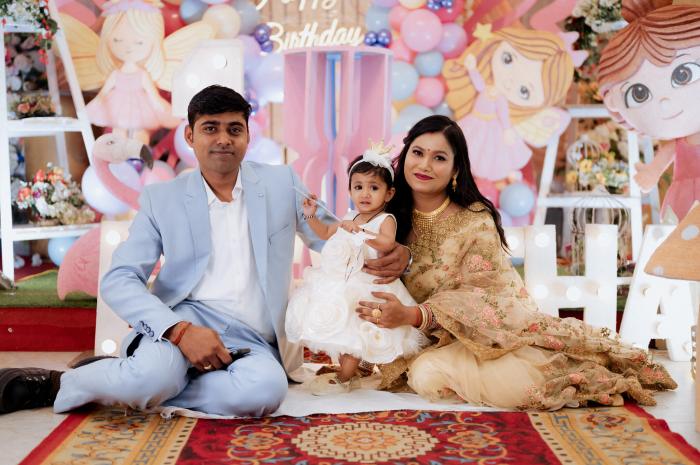 Parents and baby girl in white seated at a pink birthday backdrop, kids photography by PickYouPic Prayagraj