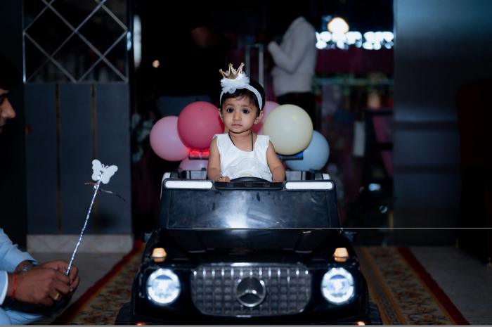 Girl wearing a crown seated in a black toy car surrounded by balloons at her birthday, by PickYouPic Prayagraj