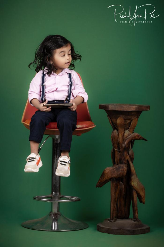 Toddler in suspenders on a studio stool against a green backdrop, kids portrait by PickYouPic Prayagraj
