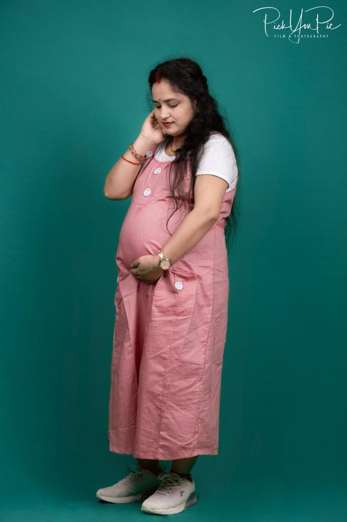 Expectant mother in a pink dungaree dress against a teal studio backdrop, maternity photoshoot by PickYouPic Prayagraj