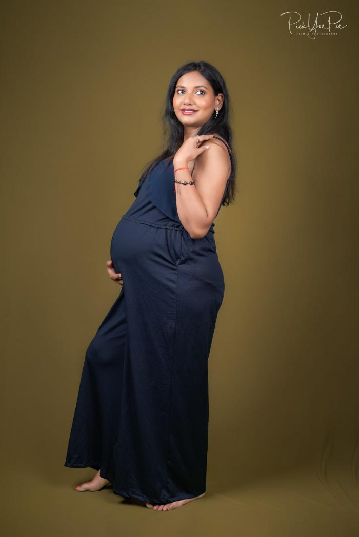 Expectant mother in a navy gown against a warm studio backdrop, maternity portrait by PickYouPic Prayagraj