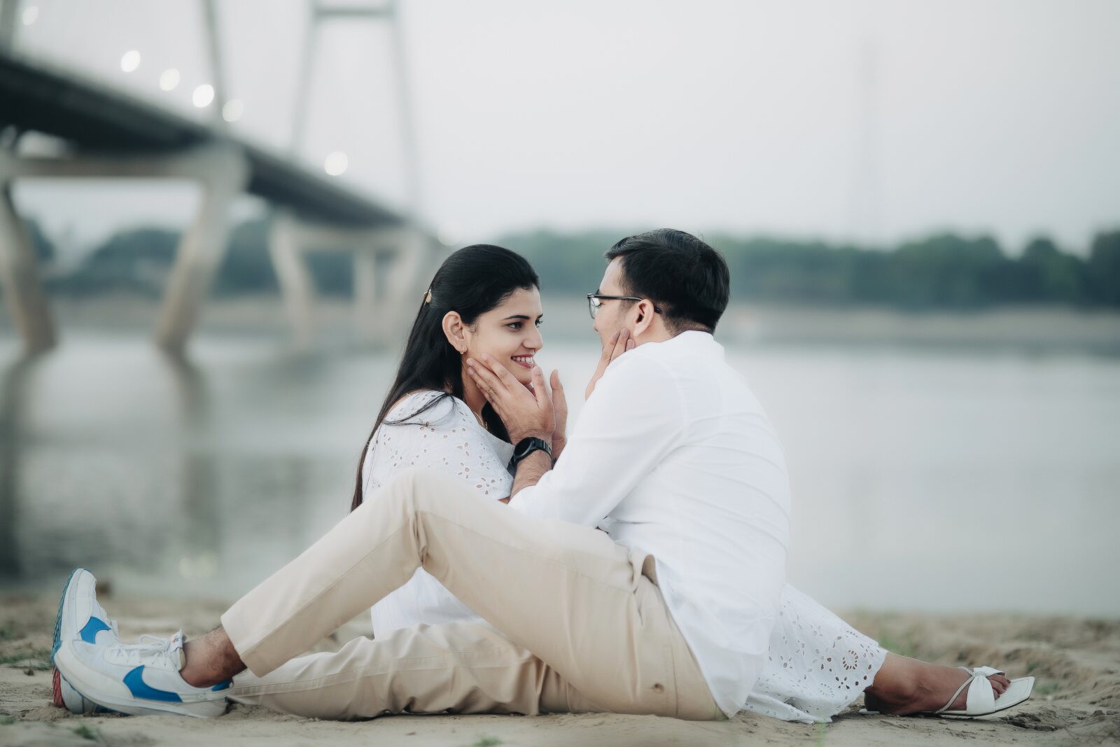 Couple sitting by the river with a bridge in the background, pre-wedding location