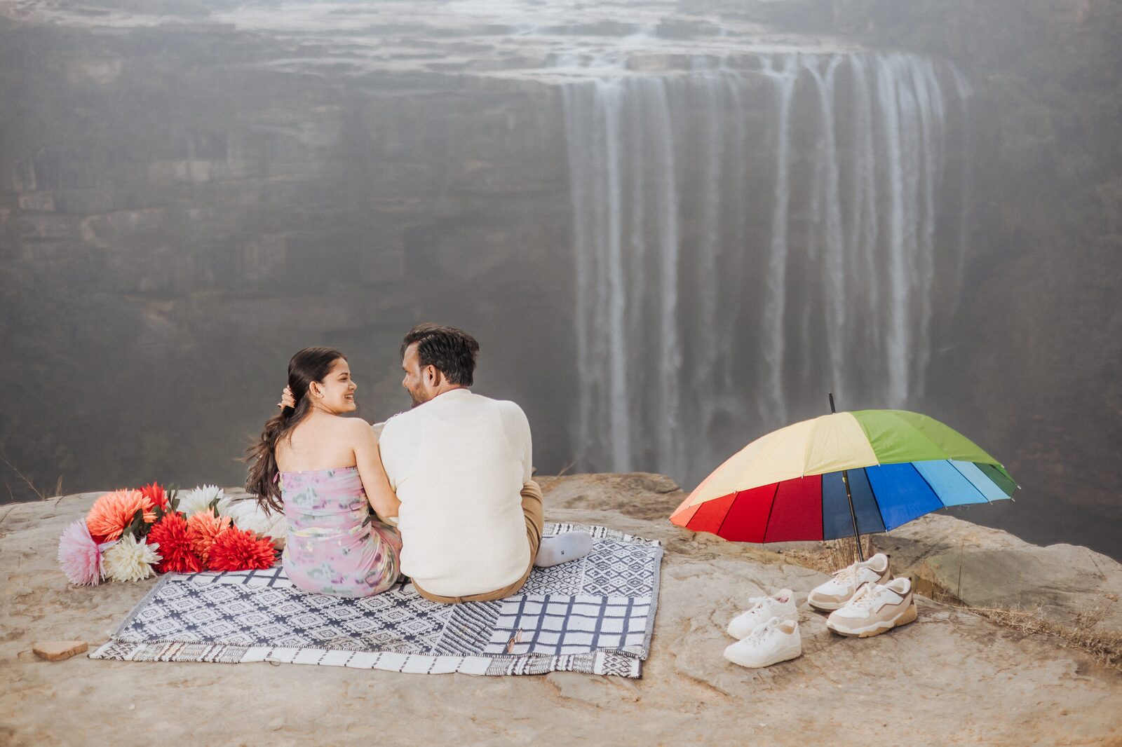 Couple at a cliff-top picnic facing Keoti Waterfall