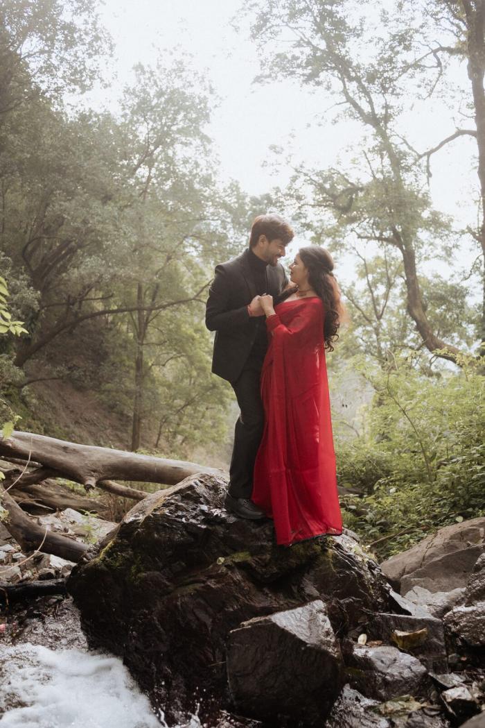 Pre-wedding shoot near a waterfall in Nainital — couple in a red saree and black suit among misty forest